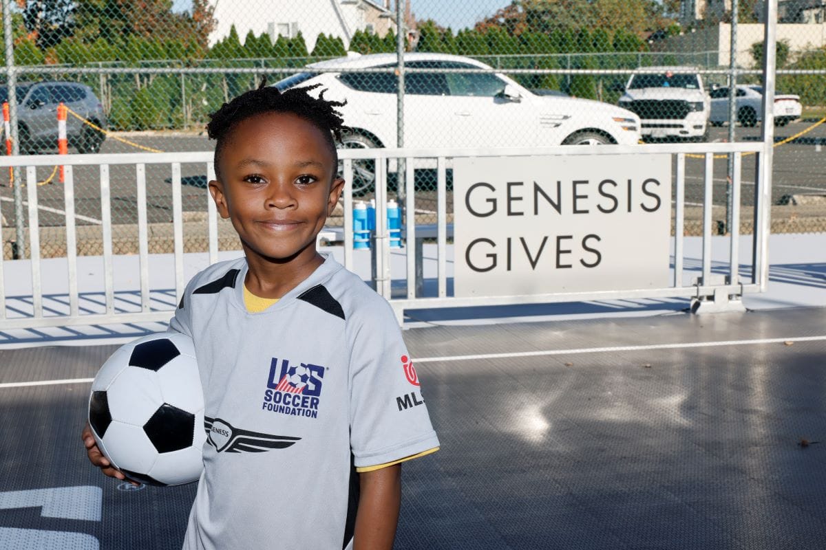 A young athlete stands in front of a sign reading "Genesis Gives" while holding a soccer ball and smiling