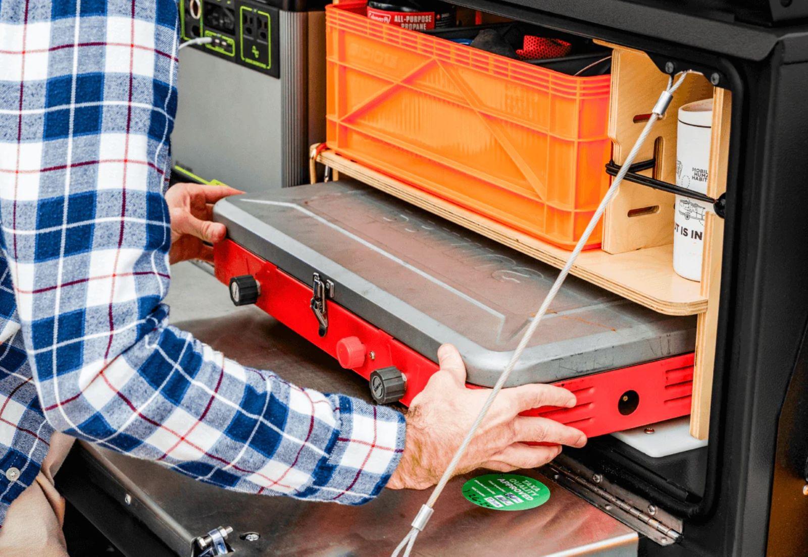A Man Pulling A Propane Powered Camping Stove From The Designated Cubby On A Taxa Woolly Bear Camping Trailer