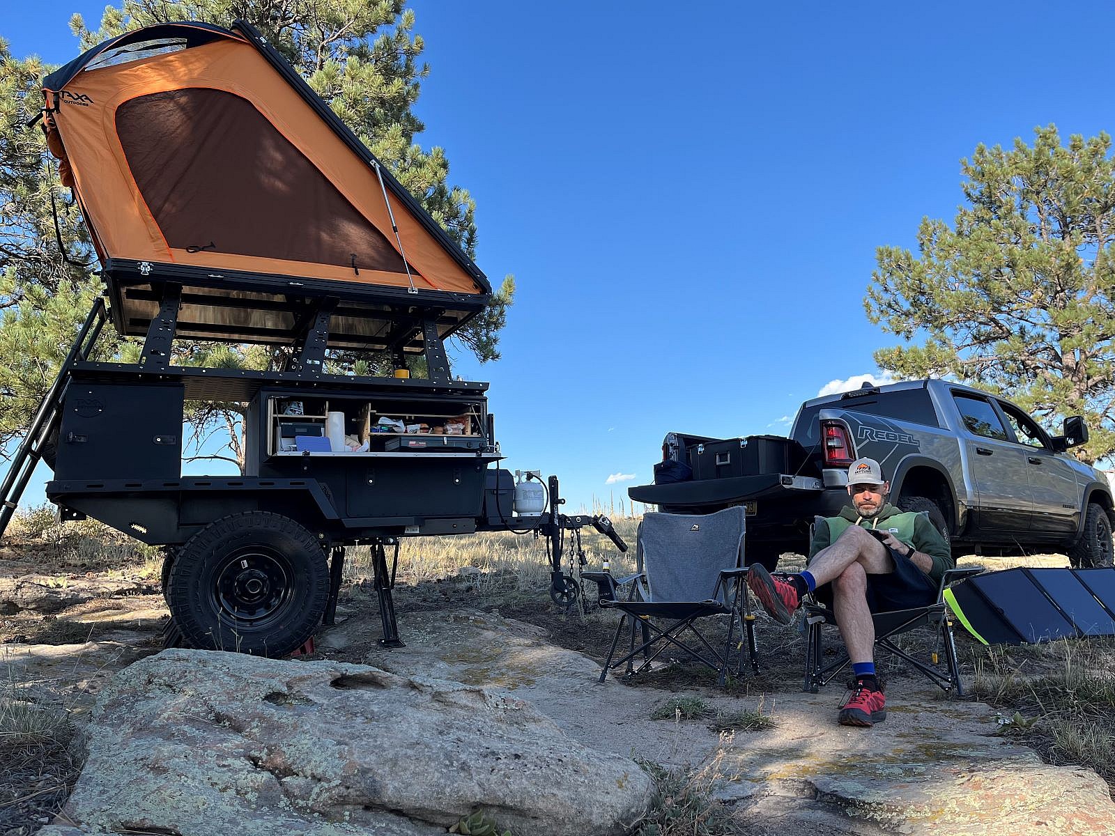 A Man Sitting At A Campsite Next To The Taxa Woolly Bear Camping Trailer Being Towed By A Large Pickup Truck