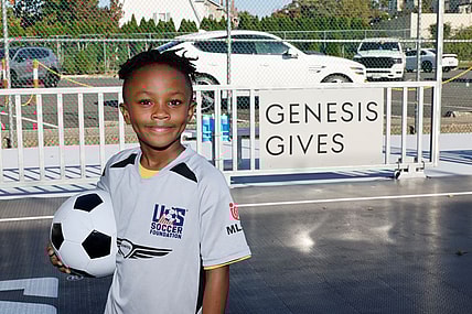 A Young Athlete Stands In Front Of A Sign Reading &Quot;Genesis Gives&Quot; While Holding A Soccer Ball And Smiling