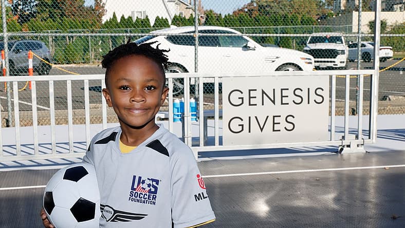 A Young Athlete Stands In Front Of A Sign Reading &Quot;Genesis Gives&Quot; While Holding A Soccer Ball And Smiling