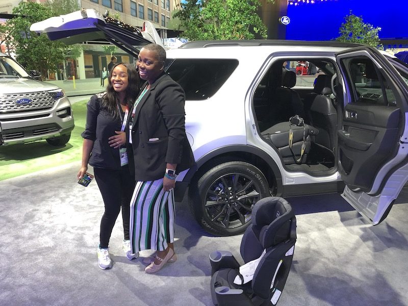 Two woman posing next to a SUV next to a child car seat